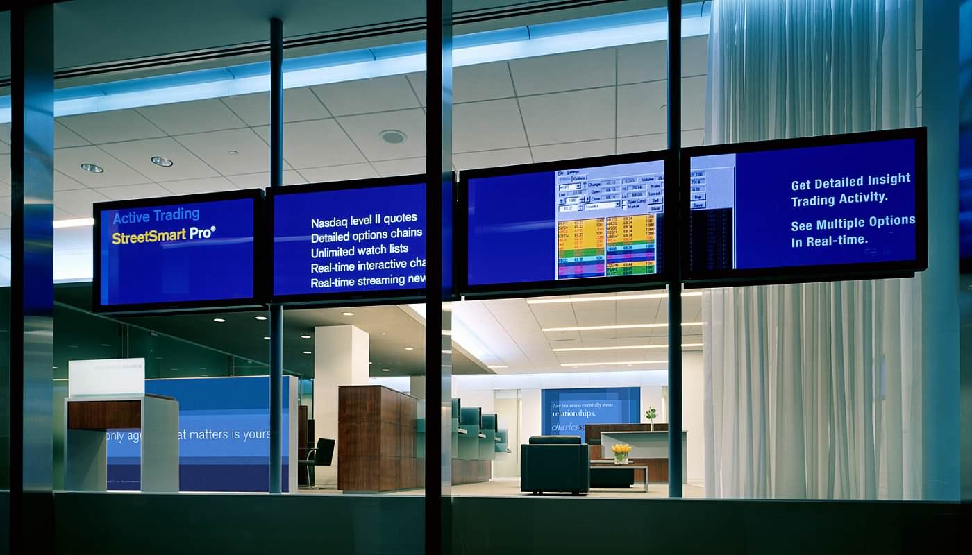 Four plasma panels mounted edge to edge behind the glass of the Charles Schwab Investor Center at night, running one sequence across all four: a title panel, a list of features, a live trading screen, and a closing line.